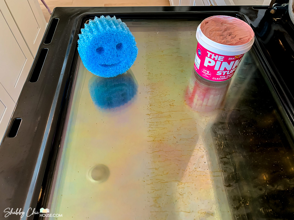 Image of a blue "Scrub Daddy" sponge and "The Pink Stuff" cleaning paste used to clean the grease from an inside of an oven window. Half of the window on the left has been cleaned and the half on the right is still full of grease.