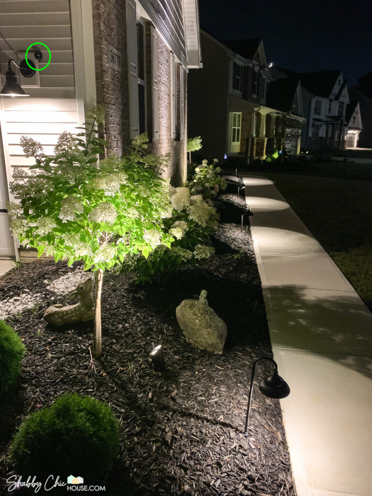Shot of landscape lighting shining on the front of a house with pathway lights, uplighting as well as a Ring Smart Lighting - Outdoor Motion sensor that when tripped can turn on all the landscape lights.