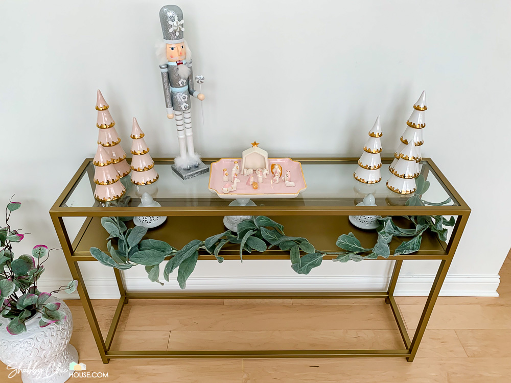 Image of a gold and glass table with two different sets of hand crafted ceramic Christmas trees. One set of trees is pink. Another set is white with 22K gold accents.
