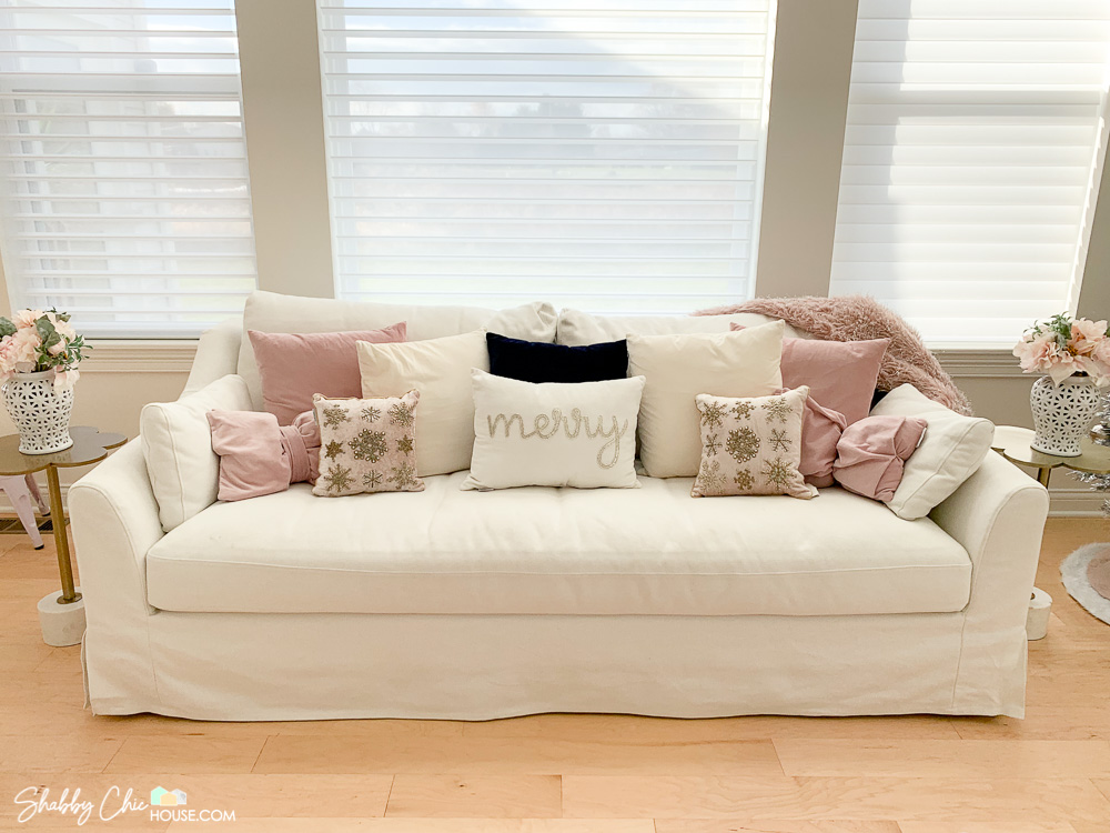 White couch with a lot of pillows. The center pillow says "Merry" and the two outer pillows are encrusted with beaded snowflakes.