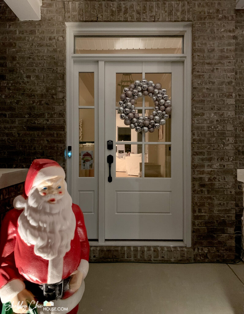 White front door with silver and pink Christmas ornament wreath with a vintage Santa blowmold in the foreground.