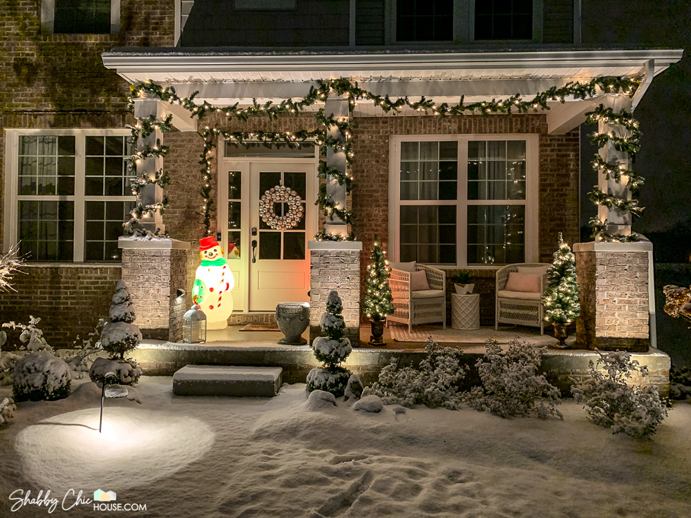 Image of a snowy front yard and front porch with Christmas decorations including pre-lit garland, two pre-lit 4' Christmas trees in planters, an ornament wreath and a Frosty the Snowman blow mold.