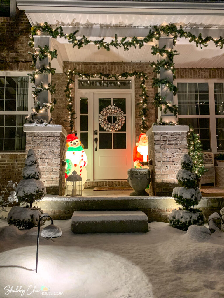 Image of a snowy front yard and front porch with Christmas decorations including pre-lit garland, an ornament wreath and a Frosty the Snowman blow mold as well as a Santa blow mold.