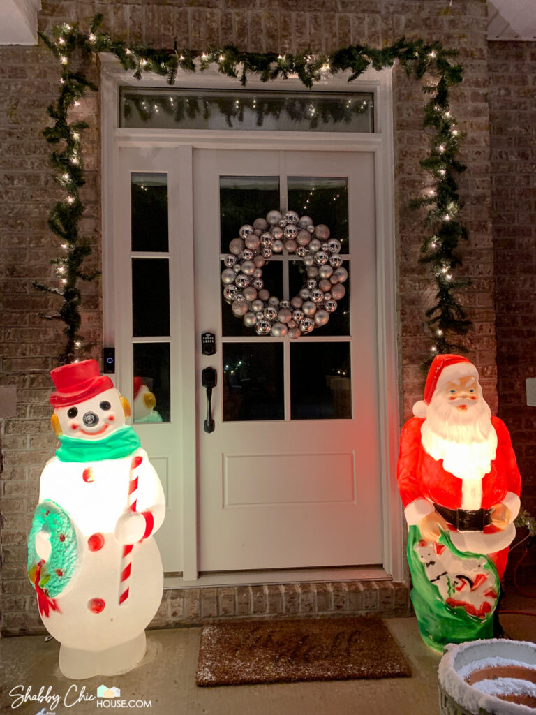 Shot of a Frosty the Snowman blow mold and Santa blow mold set-up on both sides of a front door entryway. Pre-lit garland is also wrapped around the front door and a pink and silver ornament wreath is hung on the door as well.
